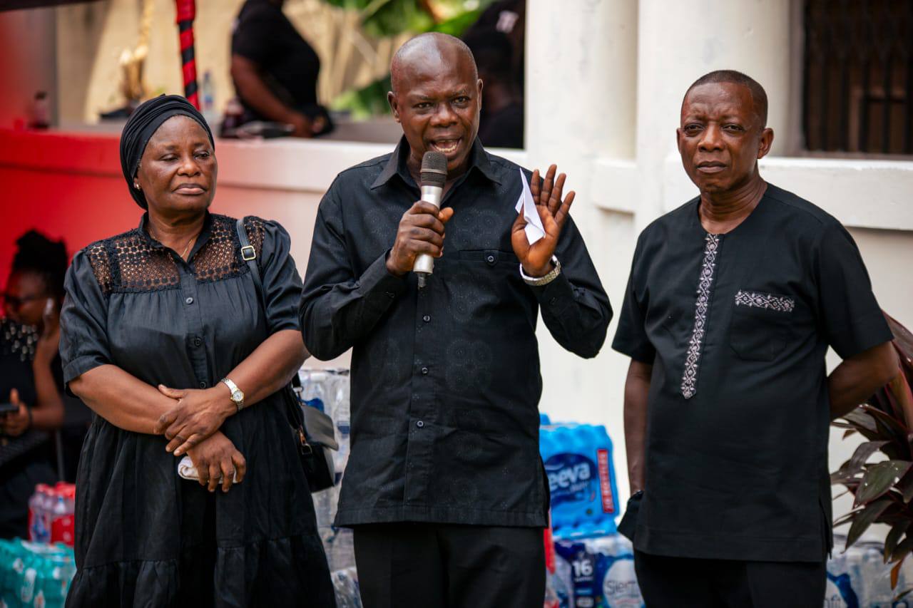The family elder and spokesperson addressing the gathered mourners from the microphone, flanked by the family matriarch and elders.